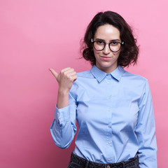 Good looking young Caucasian woman in round transparent eyewear, keeps hand raised, dressed in casual outfit, pretends holding something wonderful, isolated over pink background. Look there