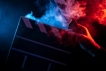 Close-up on an open clapperboard in hand before starting shooting a film with multi-colored smoke around with red and blue backlighting on a black isolated background - obrazy, fototapety, plakaty