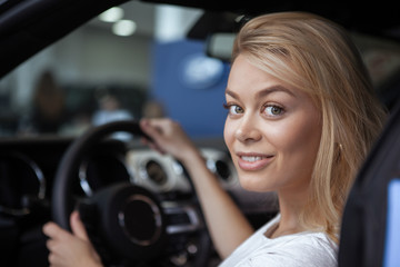 Fototapeta na wymiar Close up of beautiful blond haired woman smiling over her shoulder to the camera, sitting behind the steering wheel of a new auto. Gorgeous cheerful female enjoying buying new car. Dealership concept