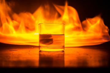 Whiskey in fire concept. Glass of whiskey and ice on wooden surface with color light and fog on background. Close up.