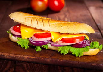 Closeup of a baguette sandwich with ham, cheese, lettuce, tomatoes and onion on a cutting board.