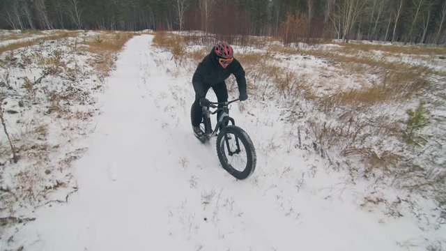 Professional extreme sportsman biker riding fat bike in outdoors. Cyclist ride in winter in snow field, forest. Man does trick on mountain bicycle with big tire in helmet, glasses. Slow motion 180fps.