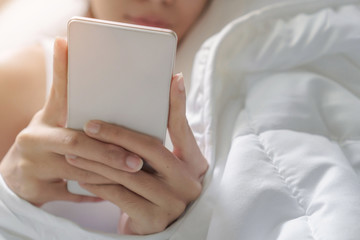 Young asian woman playing with her smathphone on white pillow on bed in bedroom on morning time after wake up, Selective focus.