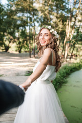 Obraz na płótnie Canvas Follow me my love concept. Attractive young woman dressed in white wedding dress holding hand of her boyfriend and walking on the beautiful garden to happy future