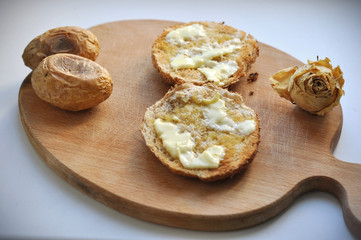 Two Rounded Slices of Bread with Butter, two baked potatoes and a dry white rose on a Wooden Chopboard.