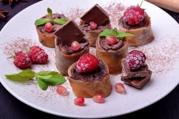 Rolled crepes with chocolate paste topped with raspberries, pomegranate, mint and chocolate pieces on a white plate