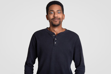 Dark skinned emotive man with bugged eyes, looks in stupor and disbelief, has stupefied facial expression, wears casual clothing, isolated over white background. People and ethnicity concept