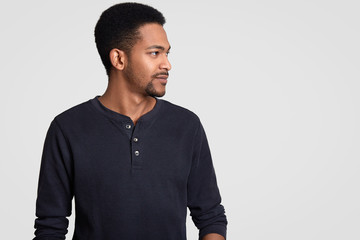 Shot of dark skinned man stands sideways at camera, notices something on free space, has serious expression, dressed in casual outfit, models over white background. Profile image of black guy indoor