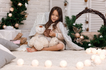 Cute girl drinks hot cocoa and plays with teddy bear at home. Christmas