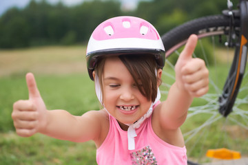 Attractive little cyclist giving thumbs up. Happy joyful little girl with bicycle on the background gesturing thumb up with two hands outdoors. Children, positive emotions and lifestyle.