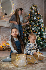 Fototapeta na wymiar Sister hugs little brother sitting on the floor with gifts on background of their parents and Christmas tree