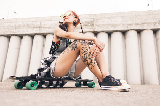 young girl with tattoo and dreadlocks sitting on a longboard on the background of concrete structure