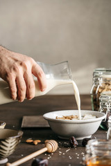 Male hand pouring milk into a white bowl with muesli, banana, honey and pumpkin seeds, Healthy morning breakfast on a wooden table