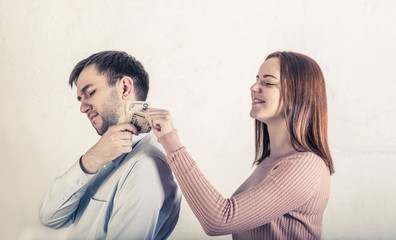Young beautiful woman taking money from dissatisfied man over shoulder on grey background. Family, husband and wife, concept of financial relations