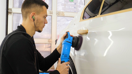 Naklejka na ściany i meble The professional guy (man) with headphones and a working uniform polishes the car body with a polishing tool. Concept from: Master class, Car Refreshment, Garage service, Special tools.