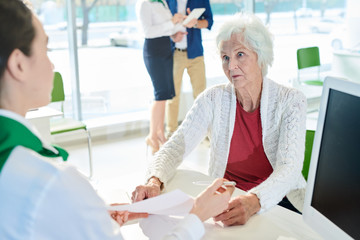 Shocked senior lady customer in white cardigan sitting at table and looking with confusion at bank...