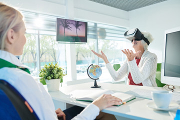 Jolly excited senior lady in cardigan sitting at table and raising hands up while using VR glasses for visualization of travel tour at meeting with trip agent.