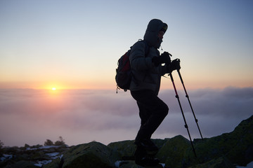 Silhouette of tourist climber in warm clothing with backpack and trekking sticks hiking on rocky mountain peak on foggy cloudy landscape, misty blue sky and raising orange sun at dawn background.