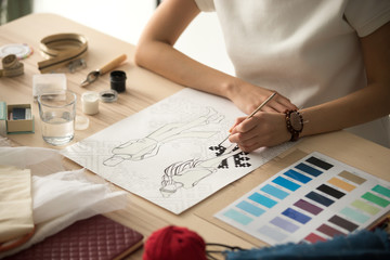 Female clothing designer coloring sketch, drawing with brush at workplace close up in workshop...