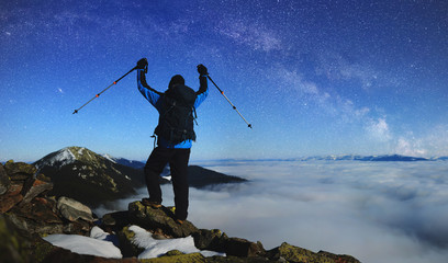 Back view of tourist hiker with backpack and trekking sticks standing with raised arms on huge rocks on background of foggy valley filled with white clouds, distant mountain tops and blue starry sky.