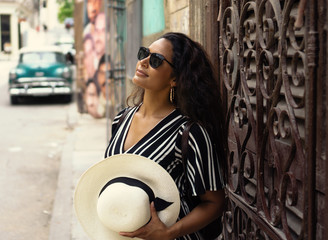Young beautiful black woman with a typical cuban hat standing in the old streets of Havana Cuba in front of a classic car.