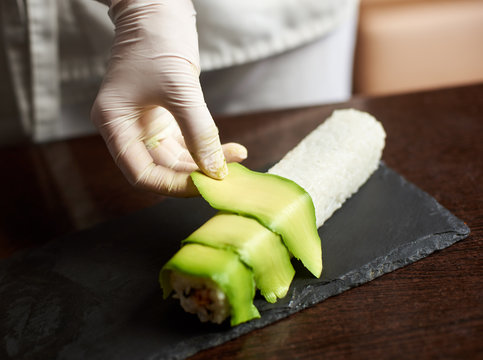 Close up view of process of preparing rolling sushi at. Hand in glove decorates roll with sliced avocado
