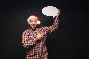 Funny serious  man in red Christmas Santa hat holds empty white cardboard as blank or mock up with copy space for text. Black background