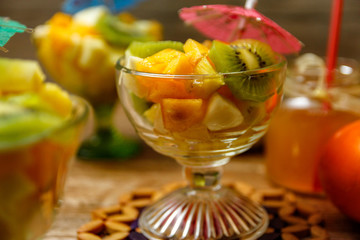 Cup of fresh fruit salad with different tropical fruit on a wooden table with raw fruits in the background. the ingredients are kiwi, persimmon, pineapple and apple