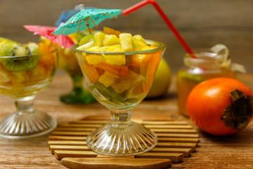 Cup of fresh fruit salad with different tropical fruit on a wooden table with raw fruits in the background. the ingredients are kiwi, persimmon, pineapple and apple