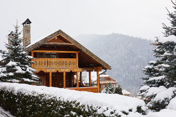 Christmas wooden mansion in mountains on snowfall winter day. Cozy chalet on ski resort near pine forest. Cottage of round timber with wooden balcony. Fir-trees covered with snow. Chimneys of stone.