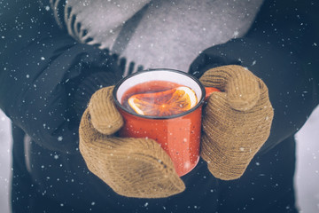 Mulled wine in a red cup in women's hands mittens. Hot winter drink outdoor in snowy winter forest....