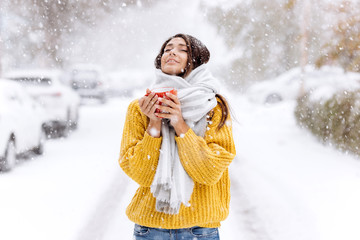 Cute dark-haired girl in a yellow sweater, jeans and a white scarf standing with a red mug on a snowy street on a winter day