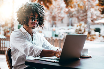 A cheerful curly Asian man freelancer in sunglasses and white shirt is chatting on the laptop with...