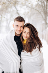 Charming dark-haired girl dressed in a yellow sweater and her boyfriend wrapped together in a gray scarf are standing in a snowy street on a winter day