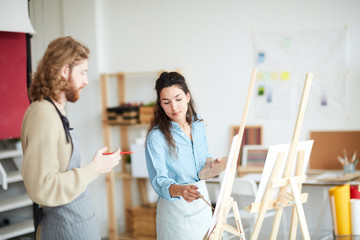 Young woman with palette pointing at her groupmate painting on easel during lesson