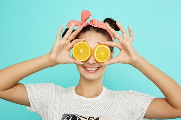 Young funny girl with pink bow on her head holds orange slices on place of her eyes on the blue background in the studio