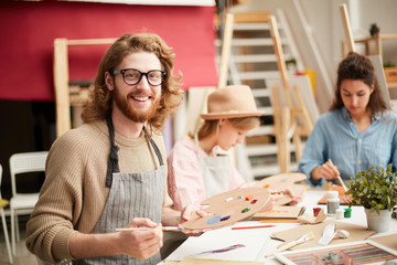 Happy young bearded man in apron looking at camera while doing painting work by table