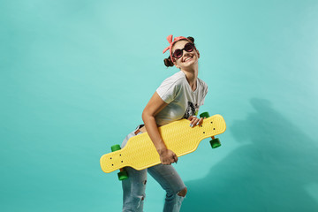 Funny young girl in sunglasses and pink bow on her head dressed in jeans and top stands with yellow...