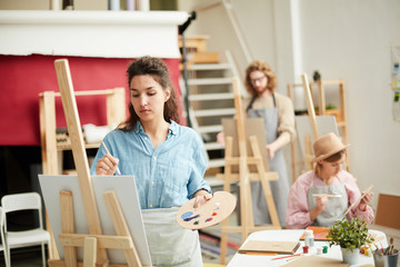 Young female in casualwear painting something on easel in modern studio of arts