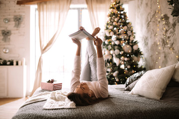 Funny girl dressed in white sweater and pants reads a book liying on the bed with gray blanket, white pillows and a New Year gift in a decorated room with a New Year tree