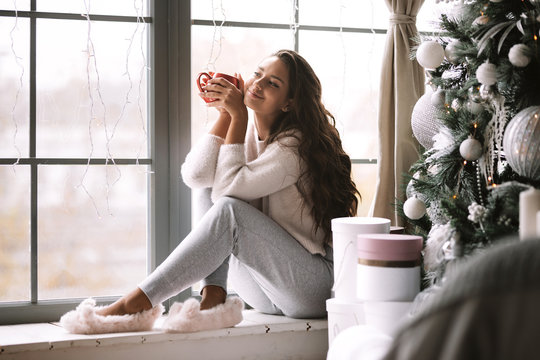 Nice dark-haired girl dressed in pants, sweater and warm slippers holds a red cup sitting on the windowsill of a panoramic window in the room next to the New Year tree and gifts
