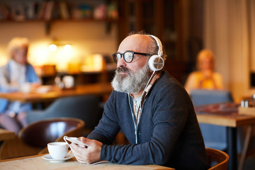 Pensive senior man with headphones and smartphone listening to relax music by table in cafe
