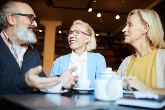 Two happy aged females listening to senior man during discussion of news by cup of tea