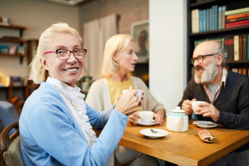 Happy blonde aged female with cup of tea sitting by table with her senior friends in cafe