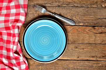 Blue ceramic plate, fork and red checkered tablecloth left frame on old vintage wooden table background - view from above