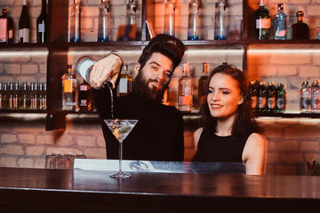 A bearded bartender with a confident look makes a cocktail with a beautiful girl behind the bar counter