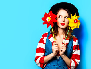Portrait of young surprised red-haired white european woman in hat and red striped shirt with jeans dress with pinwheels on blue background
