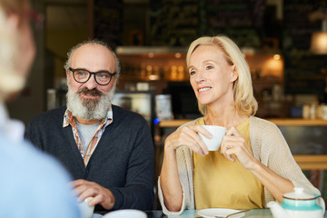 Aged casual couple with drinks looking at their friend while listening to her during conversation