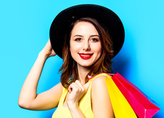 Fototapeta premium Portrait of young smiling red-haired white european woman in hat with shopping bags on blue background