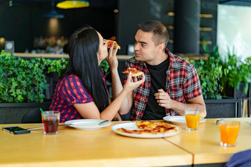 Young lovely couple is sharing pizza and eating it in pizzeria. A guy is feeding his girl with piece of pizza in a restaurant.